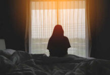 Silhouette of person sitting on bed facing window.