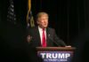 Man speaking at podium with TRUMP sign.