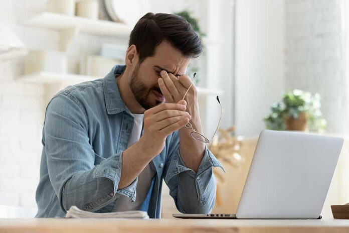 Man rubbing eyes holding glasses near laptop