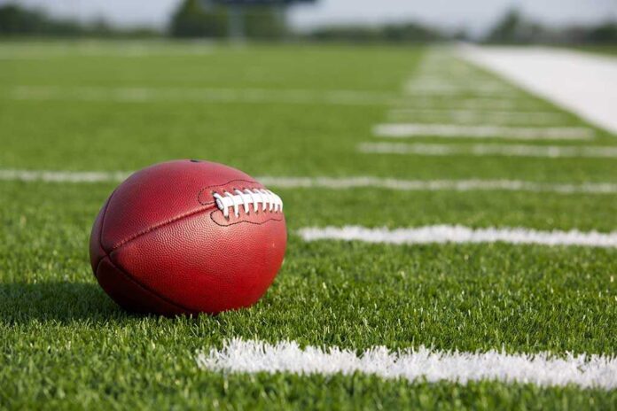 A red American football resting on a green artificial turf field