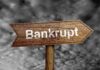 Wooden arrow sign pointing towards the word Bankrupt against a cloudy background