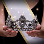 Sash-wearing person holding an ornate jeweled crown.
