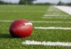 A red American football resting on a green artificial turf field
