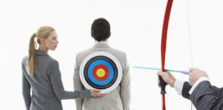 A woman places a target on a mans back while another man aims a bow and arrow at it