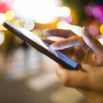 A persons hand using a smartphone in a city at night with blurred lights in the background
