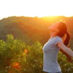 A woman standing outdoors with arms outstretched against a sunset backdrop