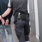 Detention Center: Force-Feeding Allegations Trigger Inquiry Officer escorting handcuffed person down hallway.
