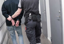 Detention Center: Force-Feeding Allegations Trigger Inquiry Officer escorting handcuffed person down hallway.