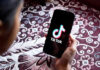 Teen Social Media Ban Sparks ID Row Hand holding phone showing TikTok logo.