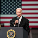 Sudden Health Fear Reopens Fitness Debate Man speaking at podium with US flag background.