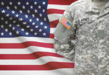 Soldier in camouflage uniform standing before American flag.