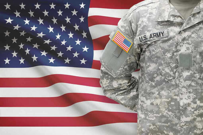 Soldier in camouflage uniform standing before American flag.
