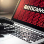 Millions Lost. Critical Systems Hit Gloved hand on laptop with ransomware screen.