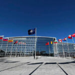 The vote is over. The debate is just beginning. Flags outside NATO headquarters building under clear blue sky.