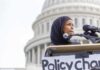 Woman in hijab speaking at podium with microphones outside a domed building