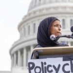 Woman in hijab speaking at podium with microphones outside a domed building