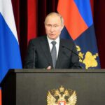 man in suit speaking at a podium with flags behind him