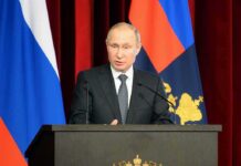Putin Has No Named Successor at 73 man in suit speaking at a podium with flags behind him