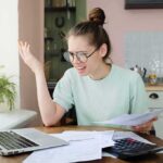 Oz Targets California Unions Over Medicaid Fraud Young woman with glasses frustrated at laptop while holding bills