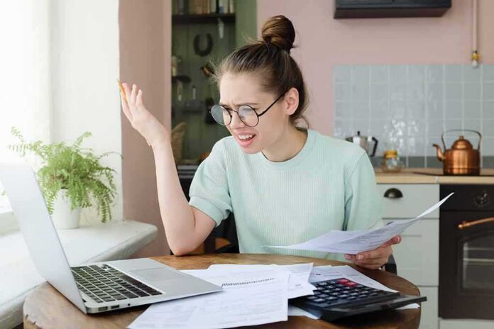 shutterstock_1119919928 Young woman with glasses frustrated at laptop while holding bills