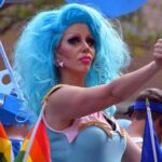 7 Days to End Secret Gender Transitions Drag performer with bright blue wig giving thumbs up at parade