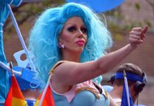 Drag performer with bright blue wig giving thumbs up at parade