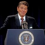 man at presidential podium gesturing with arms open