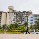 Partially collapsed beachfront apartment building behind people walking on sand