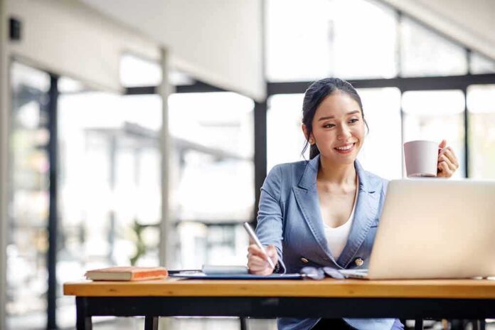 shutterstock_2149123217-1 Woman working on a laptop while holding a mug