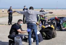 Border Emergency Declared After Migrant Wave Police officers process group of people on a beachside pavement