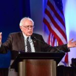 elderly man speaking at a podium with arms outstretched