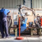 Billions Bet On Robot Wars Factory worker operates near an industrial robot arm in a workshop