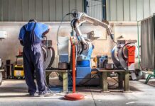 Billions Bet On Robot Wars Factory worker operates near an industrial robot arm in a workshop