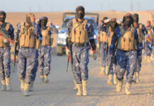 Camouflaged soldiers wearing masks and vests walk in desert.
