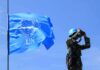 Trump’s “Annihilate Iran” Remark Draws Attention UN peacekeeper with blue helmet looks through binoculars beside UN flag