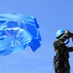 Trump’s “Annihilate Iran” Remark Draws Attention UN peacekeeper with blue helmet looks through binoculars beside UN flag