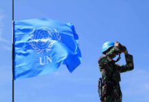 UN peacekeeper with blue helmet looks through binoculars beside UN flag