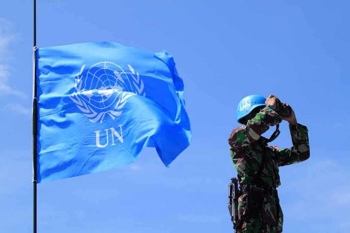 shutterstock_1480711058 UN peacekeeper with blue helmet looks through binoculars beside UN flag