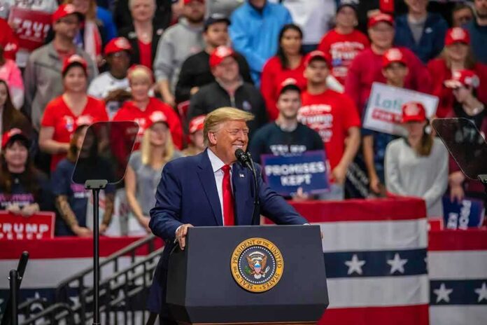Donald Trump speaks at a rally podium; Christian Jones and Bear Pascoe are in the crowd behind him